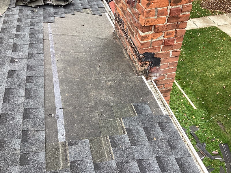 Flat roof junction with a brick wall, shingle patterns visible, partial lawn in background by Performance Roofing and Siding.