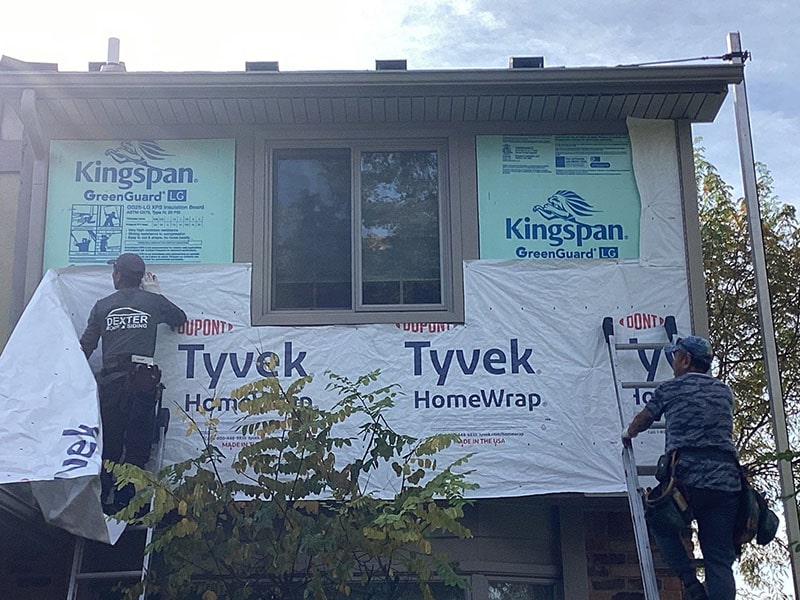 Two workers installing house wrap on the exterior of a building with insulation advertising service by Performance Roofing and Siding.