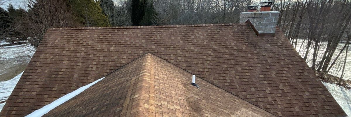 Dimensional asphalt shingles in shades of brown covering a residential roof, with a gray stone chimney and snow in the background by roof replacement companies in Michigan