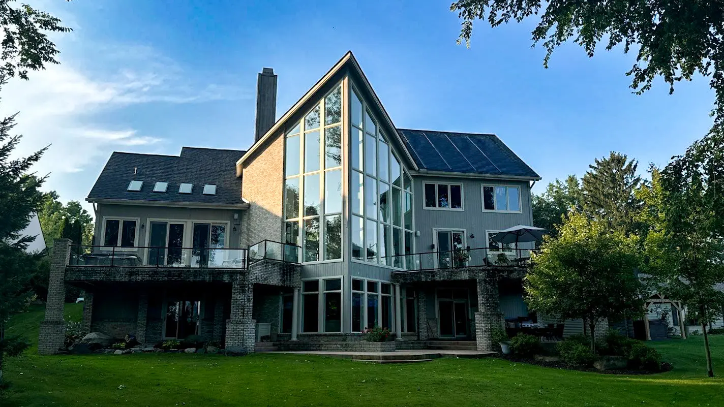 Large modern home with expansive windows and solar shingles installed on the upper roof section by Performance Roofing and Siding, surrounded by trees and lawn